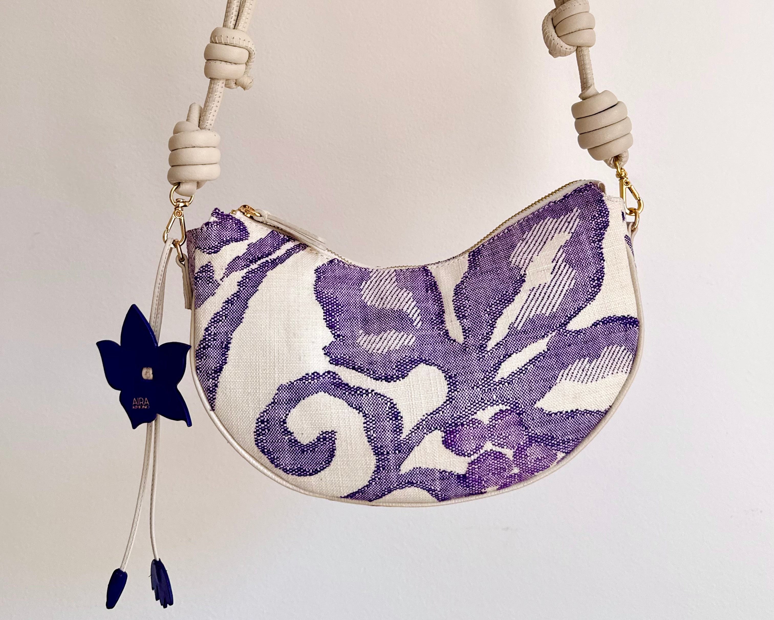 An upcycled handbag made from Japanese woven Obi sash belt,  a cream-colored knotted shoulder adjustable strap and a white body featuring a purple floral ikat pattern, and a flower bag charm, that can transform from shoulder bag, crossbody bag or clutch.
