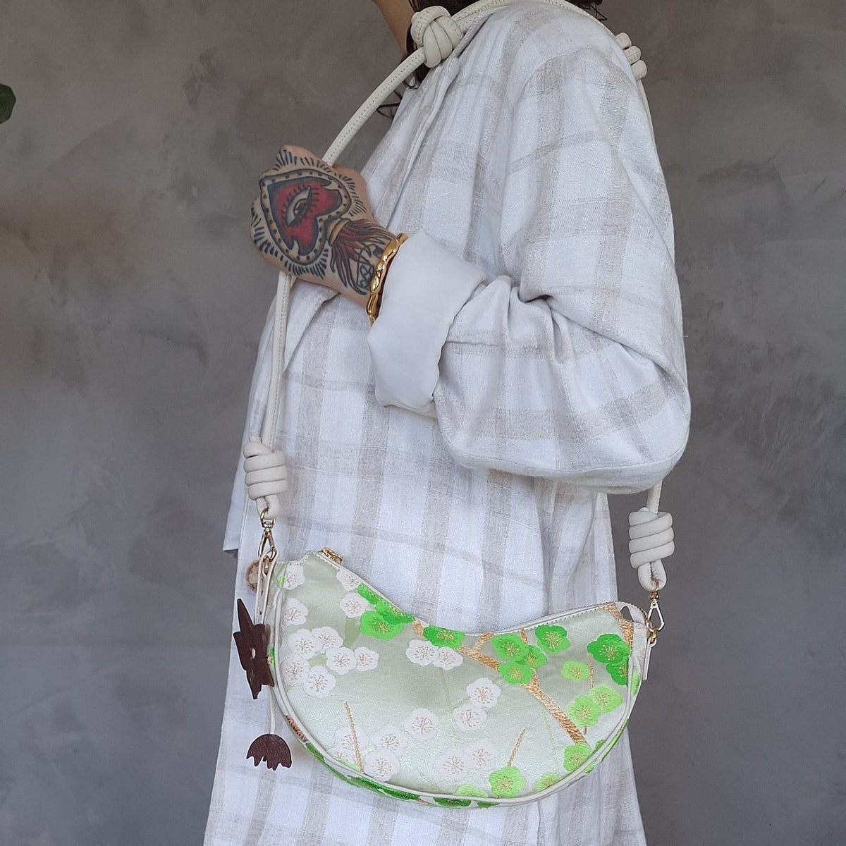 A person carries a white crescent-shaped shoulder bag or crossbody bag, with leather strap and green cherry-blossom embroidered pattern made from secondhand Japanese Obi sash and a brown flower bag-charm.