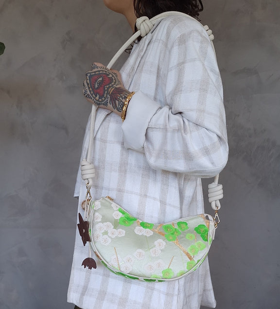 A person carries a white crescent-shaped shoulder bag or crossbody bag, with leather strap and green cherry-blossom embroidered pattern made from secondhand Japanese Obi sash and a brown flower bag-charm.