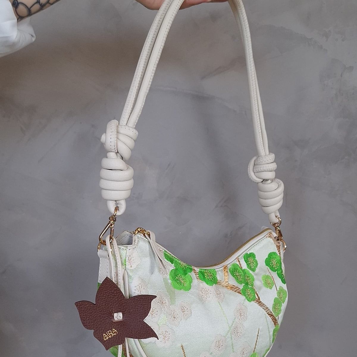 A hand holding a small crescent-shaped shoulder bag with a cream and white floral print, green leaf motifs, a knotted rope handle, and a dark brown hanging flower charm against a gray textured wall.