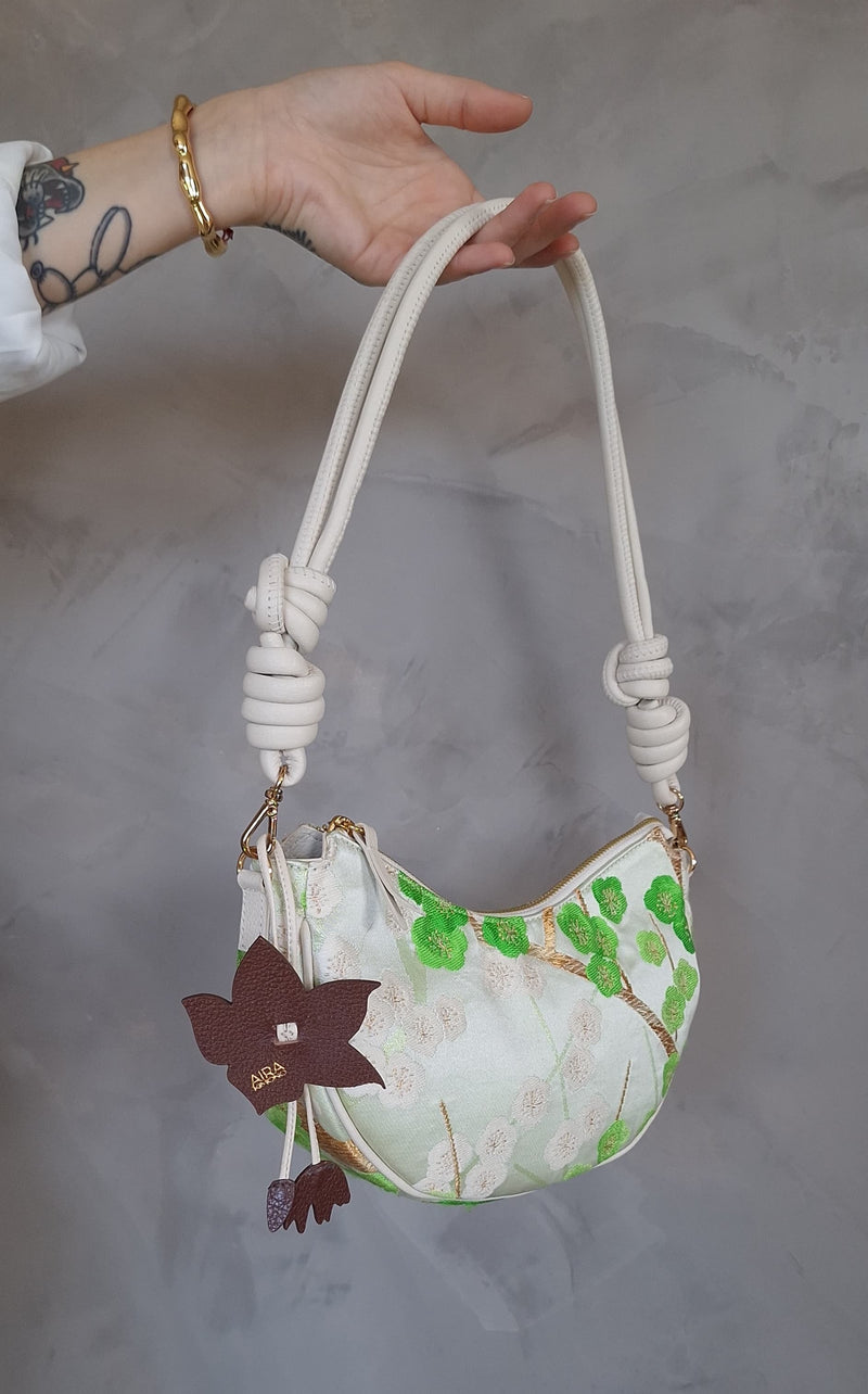 A hand holding a small crescent-shaped shoulder bag with a cream and white floral print, green leaf motifs, a knotted rope handle, and a dark brown hanging flower charm against a gray textured wall.