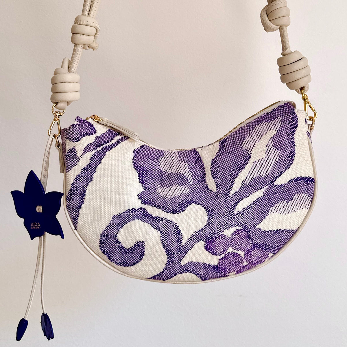An upcycled handbag made from Japanese woven Obi sash belt,  a cream-colored knotted shoulder adjustable strap and a white body featuring a purple floral ikat pattern, and a flower bag charm, that can transform from shoulder bag, crossbody bag or clutch.
