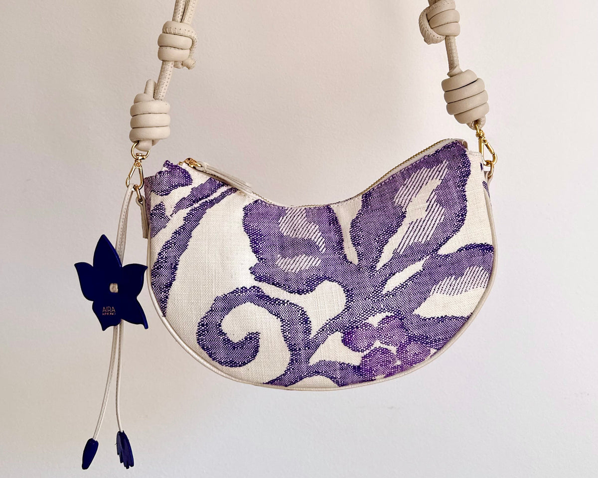 An upcycled handbag made from Japanese woven Obi sash belt,  a cream-colored knotted shoulder adjustable strap and a white body featuring a purple floral ikat pattern, and a flower bag charm, that can transform from shoulder bag, crossbody bag or clutch.
