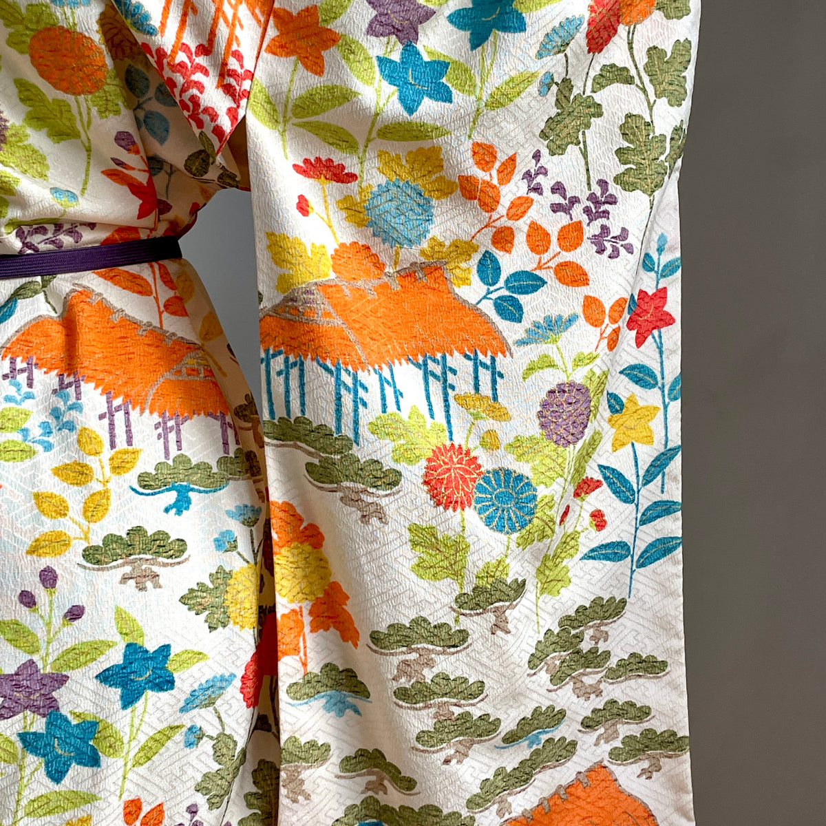 A close-up of a kimono-style garment with a cream base and a dense multicolor floral and leaf pattern, including orange, blue, green, yellow, and purple motifs. A dark purple belt is visible at the waist.