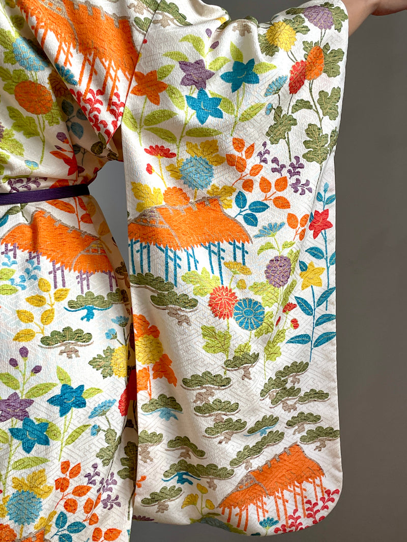 A close-up of a kimono-style garment with a cream base and a dense multicolor floral and leaf pattern, including orange, blue, green, yellow, and purple motifs. A dark purple belt is visible at the waist.