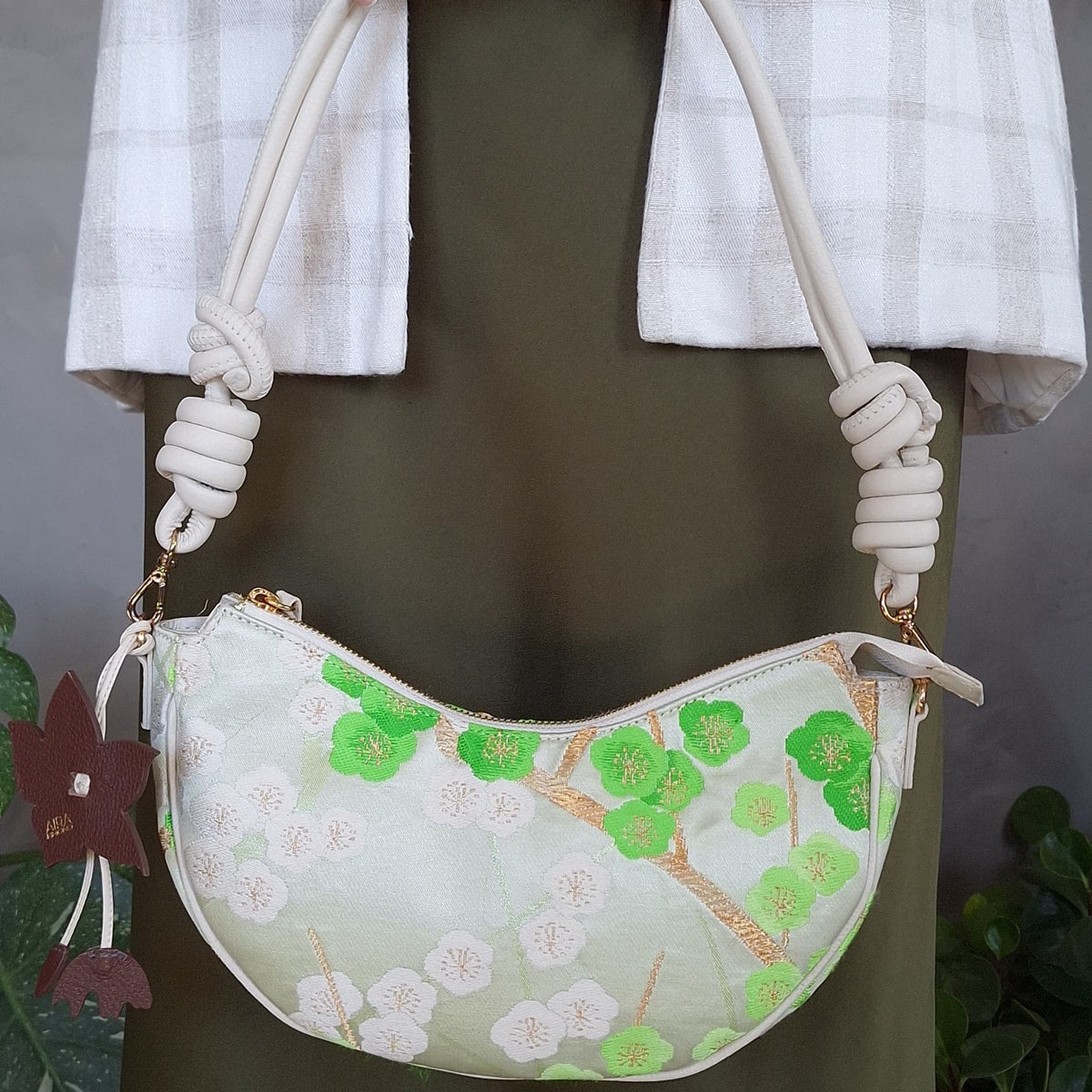 A person carries a white crescent-shaped shoulder bag or crossbody bag, with leather strap and green cherry-blossom embroidered pattern made from secondhand Japanese Obi sash and a brown flower bag-charm.