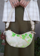 A person carries a white crescent-shaped shoulder bag or crossbody bag, with leather strap and green cherry-blossom embroidered pattern made from secondhand Japanese Obi sash and a brown flower bag-charm.