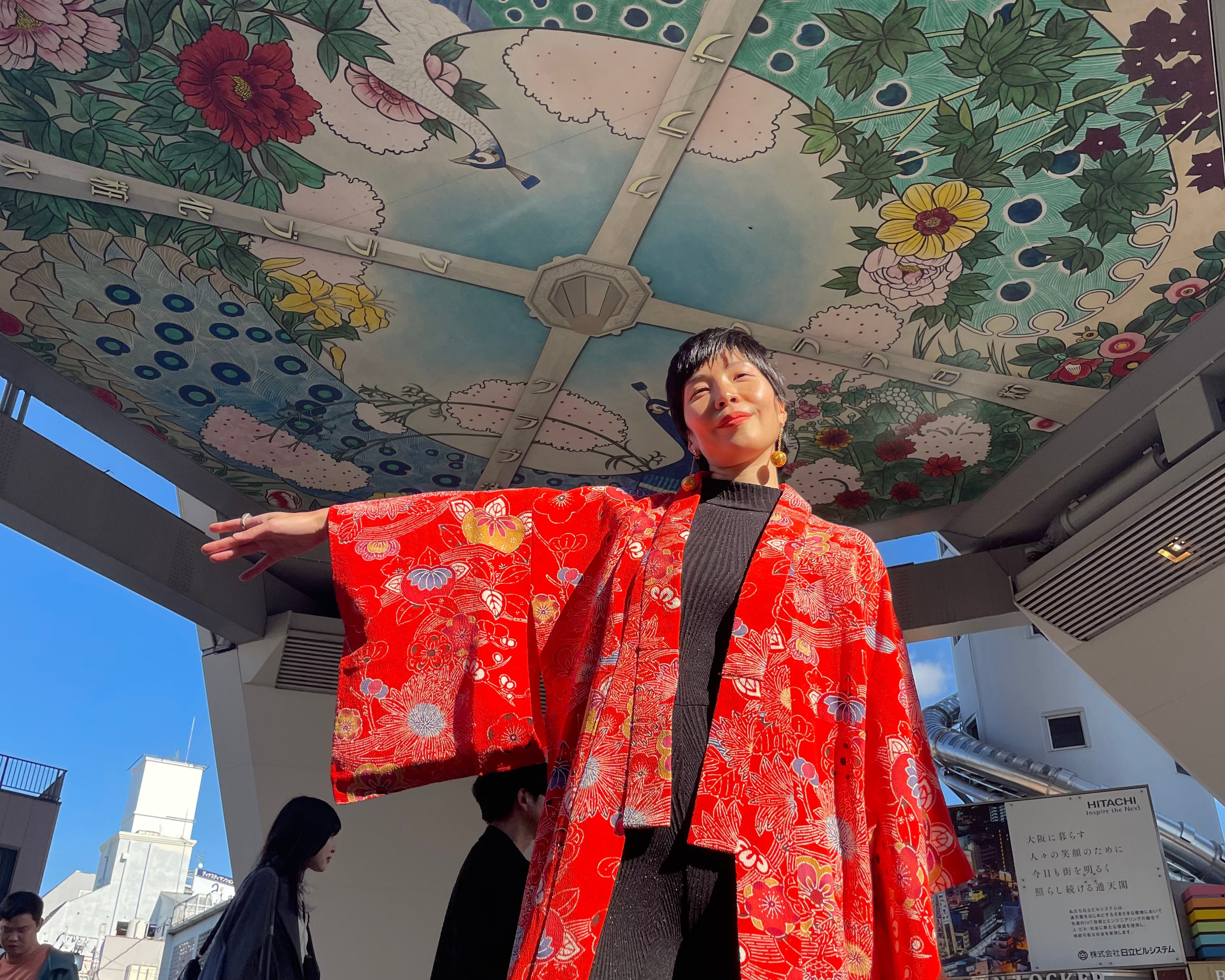A Japanese girl wearing a bright red kimono Bingata from Japan, with a white floral pattern and a black top underneath, standing under a large decorative ceiling in Osaka, with colorful painted floral and bird motifs in an outdoor urban setting.